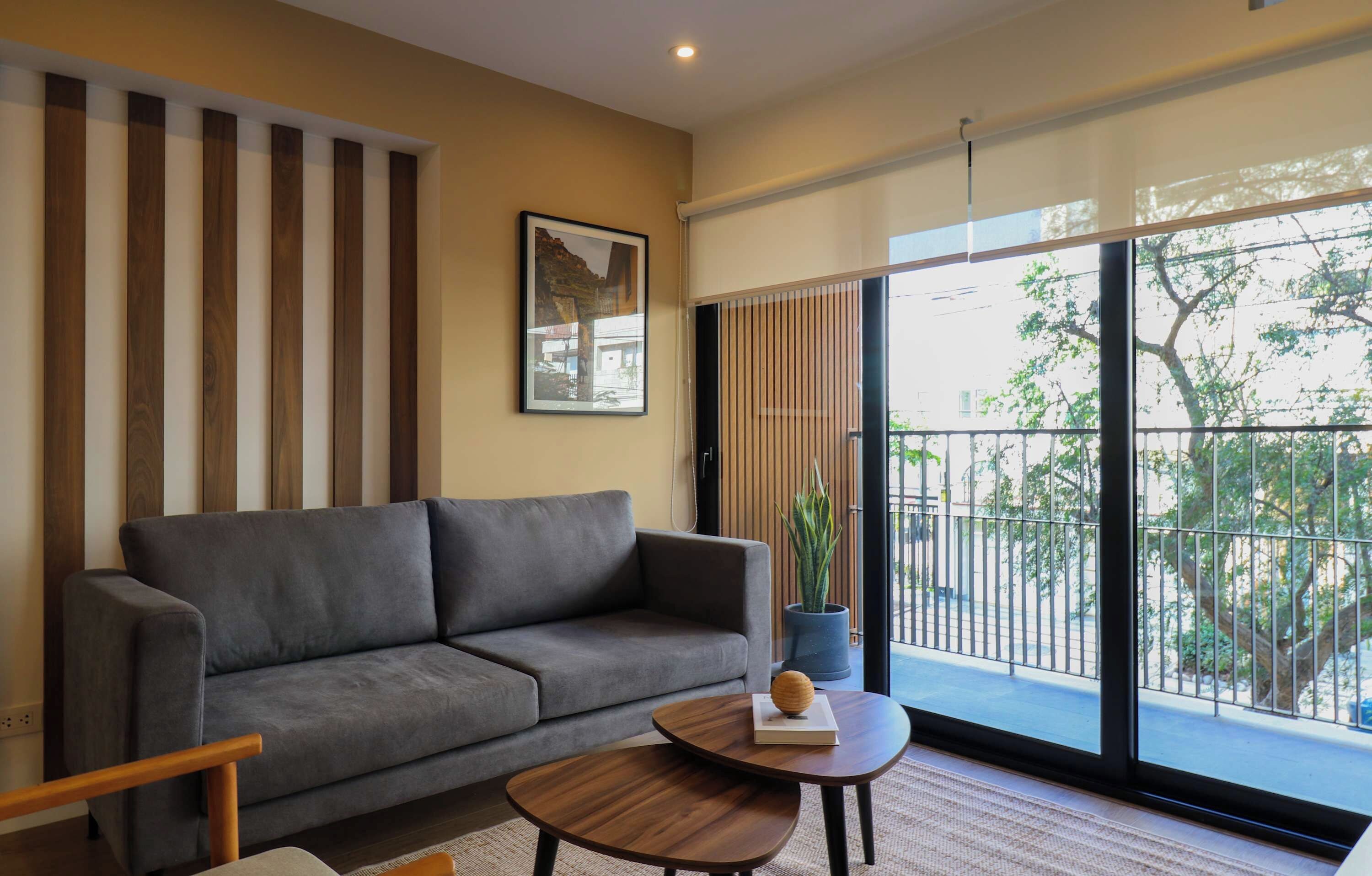 A modern living room with a grey sofa, a wooden coffee table, and a large window with a view of trees and buildings.