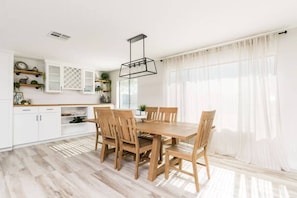 Eine moderne Küche mit weißen Schränken, einem Holzespeisetisch und Stühlen. Ein Fenster mit durchscheinenden Vorhängen und eine Pendelleuchte über dem Tisch.
