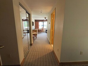 Un pasillo de hotel con piso alfombrado, un comedor con mesas y sillas, y vista al exterior a través de amplias ventanas.