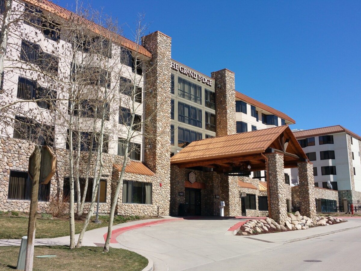 Un hotel de varios pisos con fachada de piedra, un acceso cubierto y un cielo azul despejado.