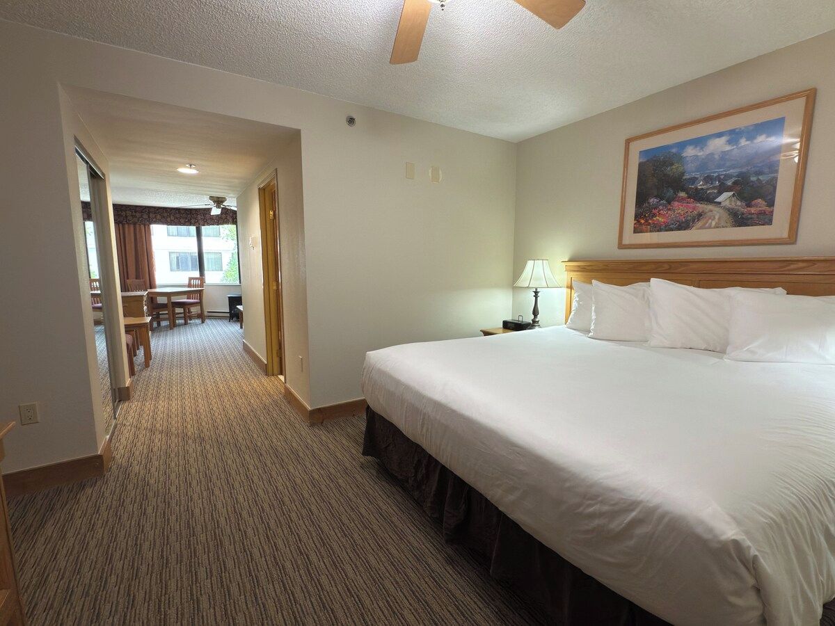 A hotel room with a large bed, a desk, and a chair. There is a painting on the wall and a window with curtains.