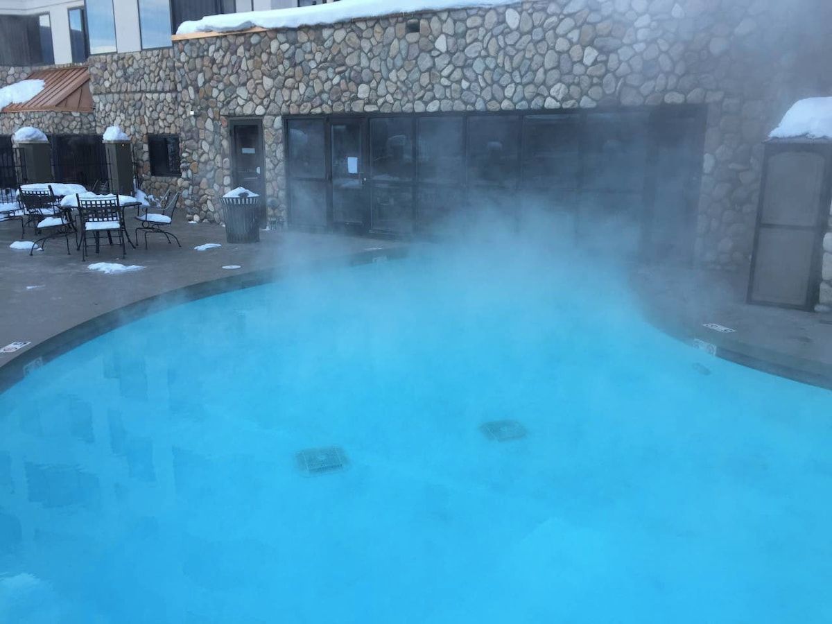 Edificio de piedra con piscina que emite vapor, rodeado de mobiliario de exterior.