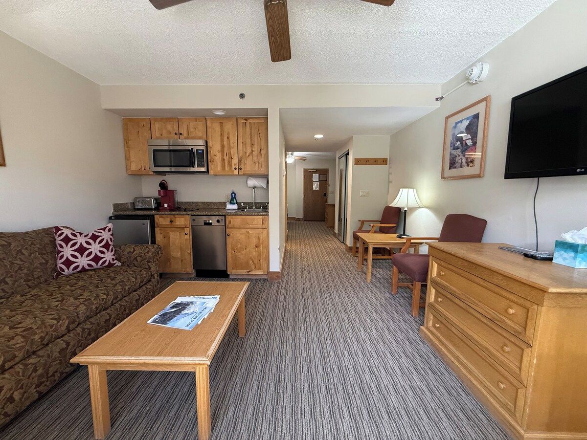 A compact living space with a kitchenette, a sofa, a coffee table, a TV, and a desk with drawers.