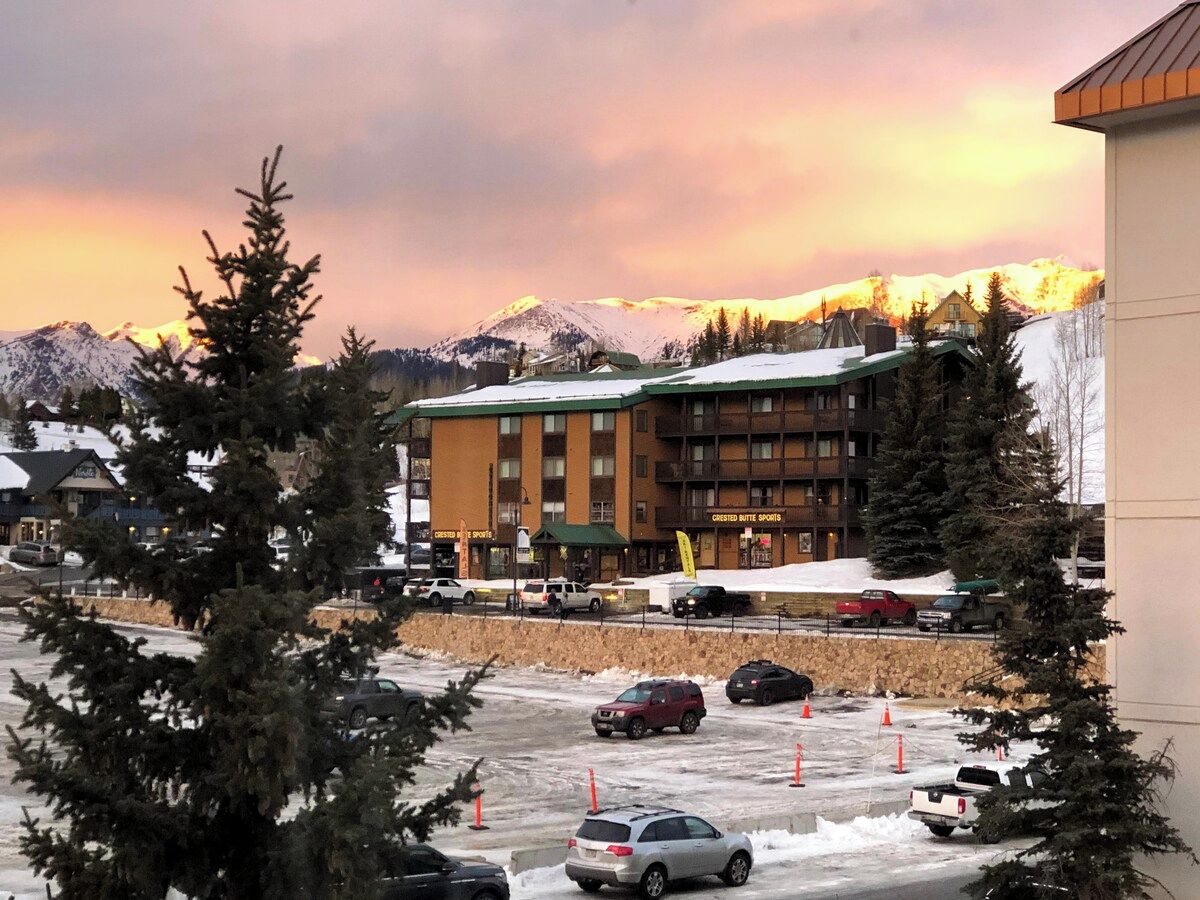 A hotel with a parking lot, snow-covered mountains in the background, and a sunset sky.
