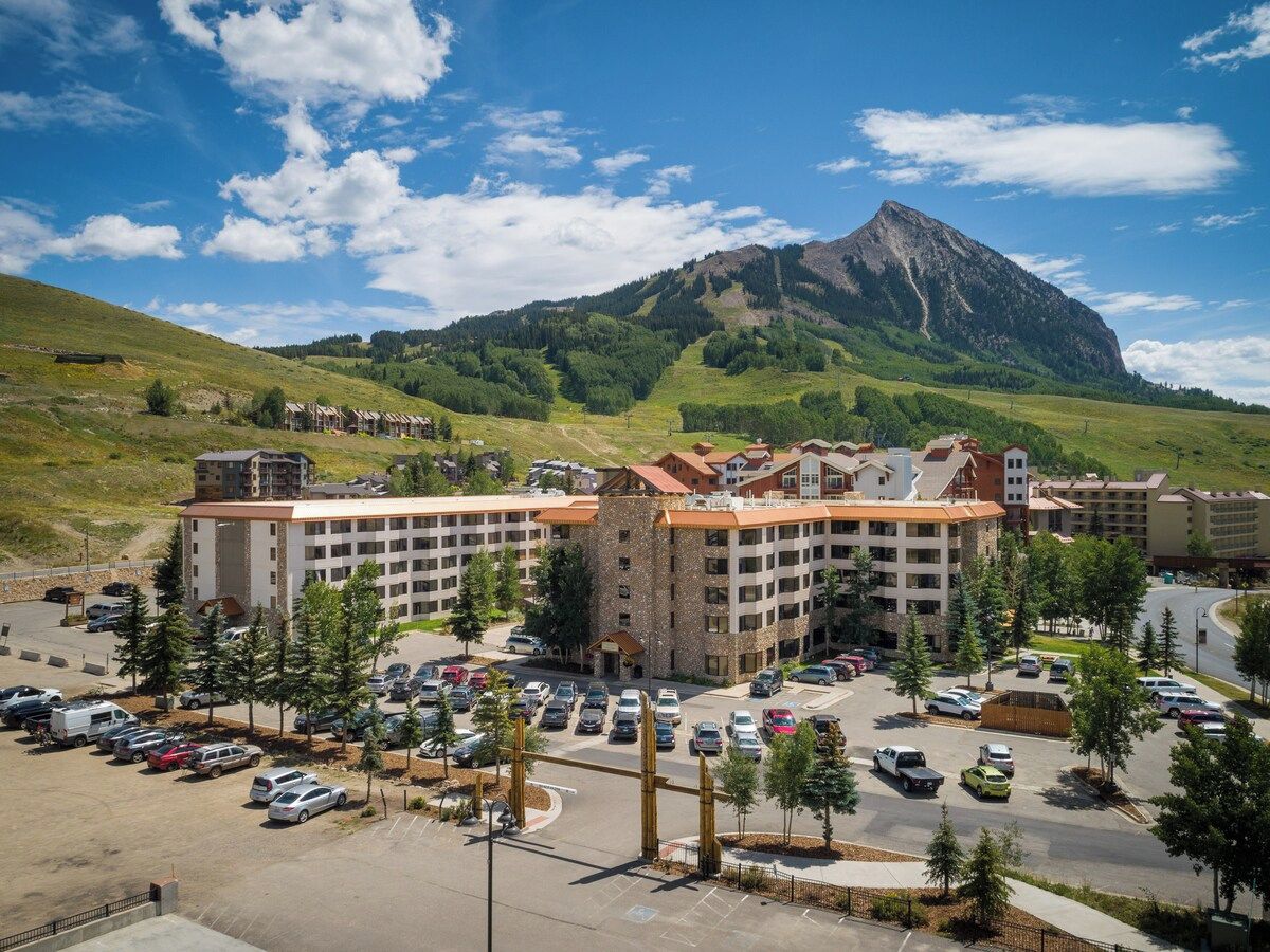 A multi-story hotel with a parking lot, surrounded by greenery and a mountain in the background.