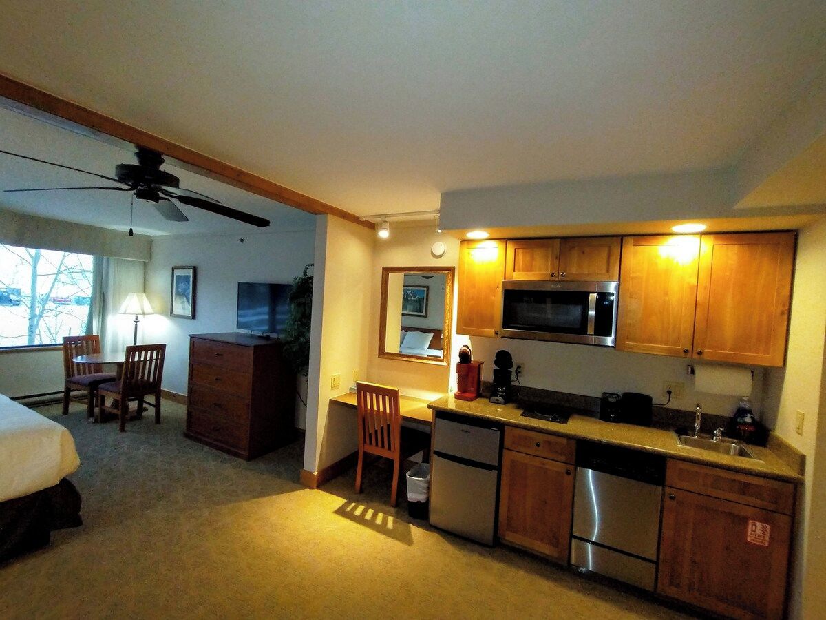 A hotel room with a kitchenette, a bed, a desk, a chair, and a ceiling fan.