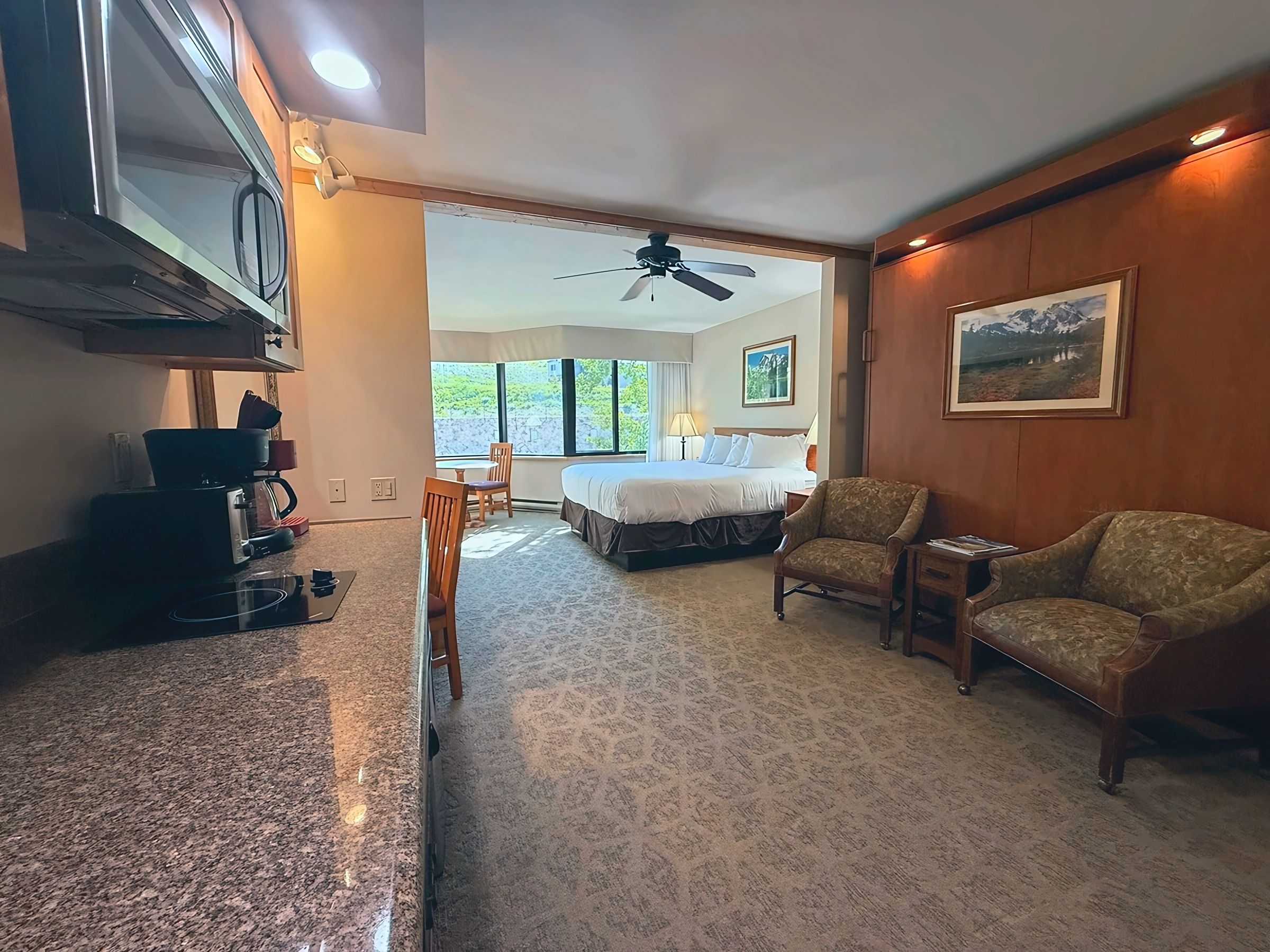 A hotel room with a bed, two chairs, a dining table, a coffee maker, and a view of the outdoors.
