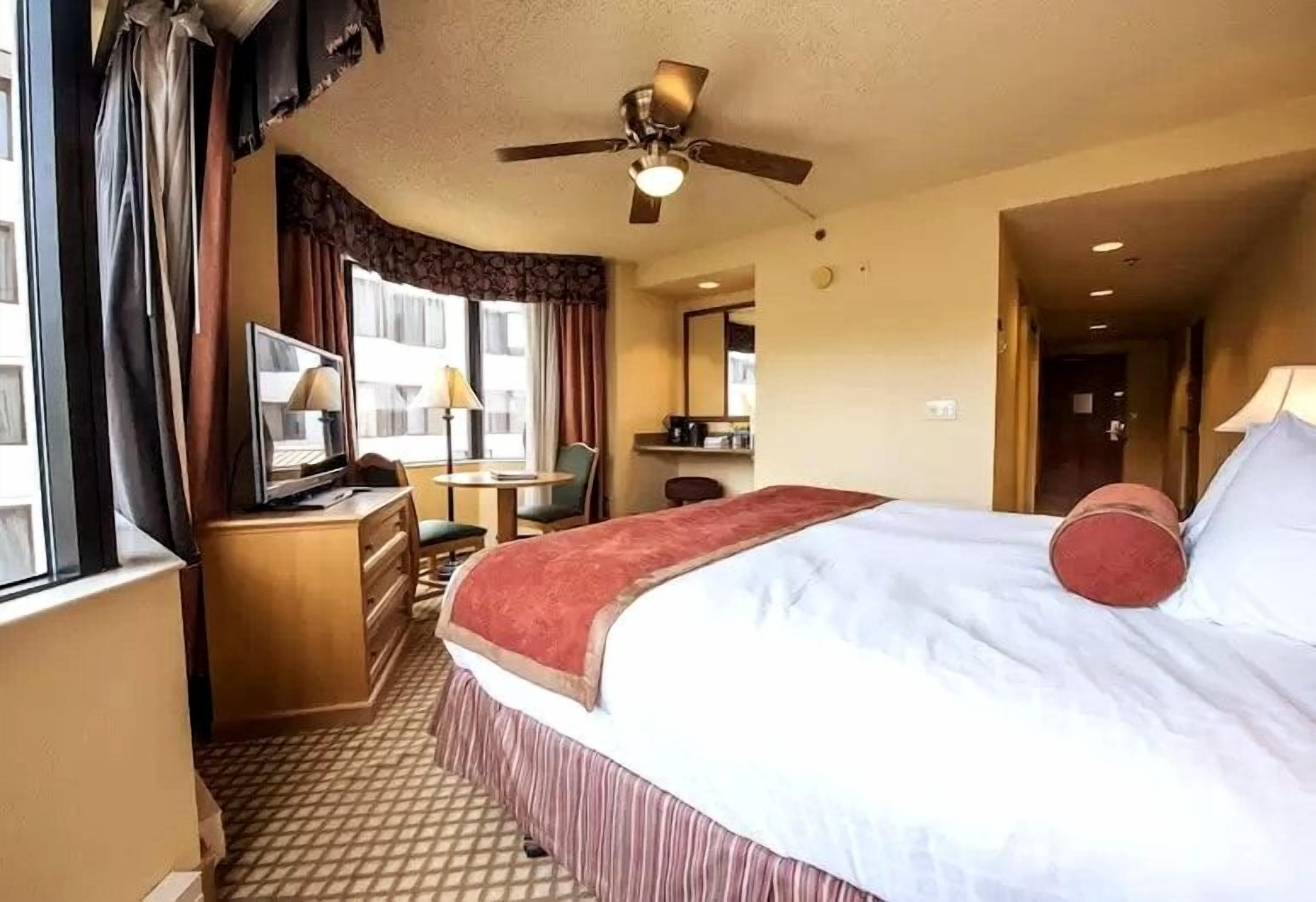 A hotel room with a bed, a desk with a computer, a chair, a ceiling fan, and a window with curtains.