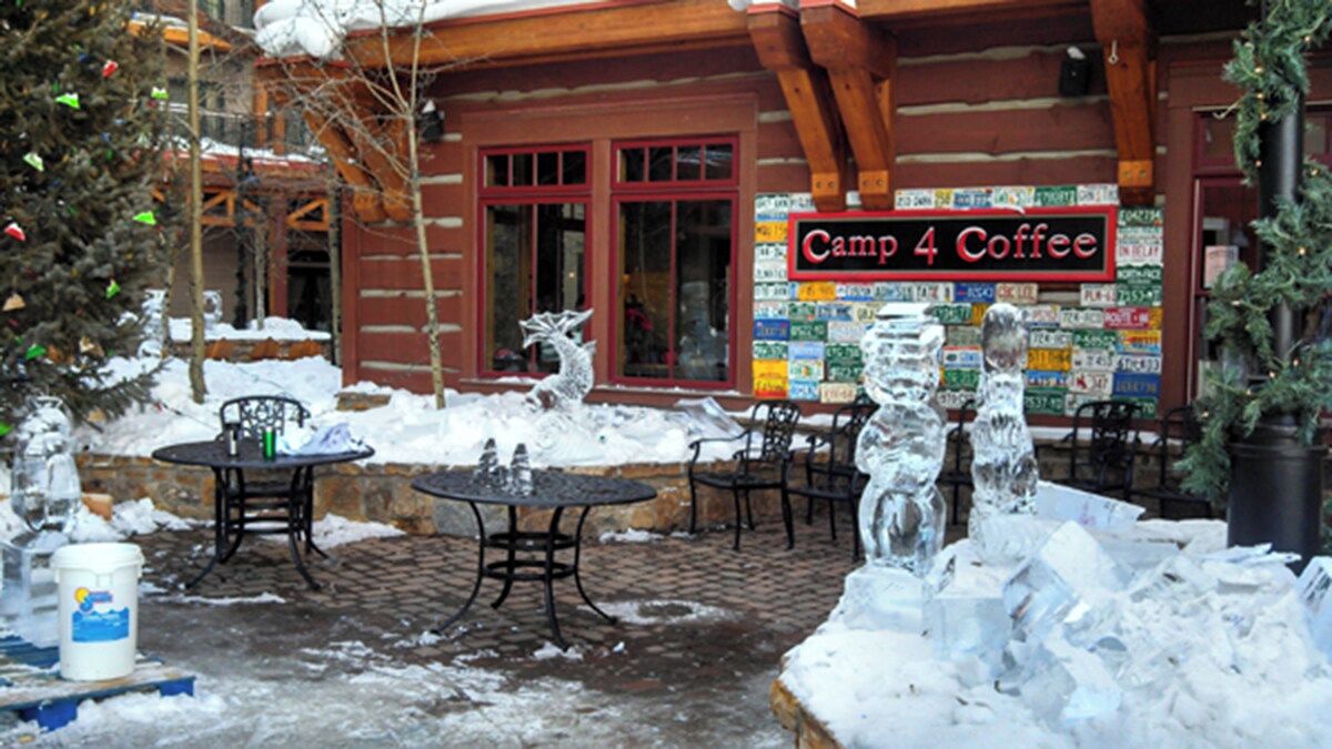 A wooden building with a sign reading 'Camp 4 Coffee' and outdoor seating with tables and chairs.