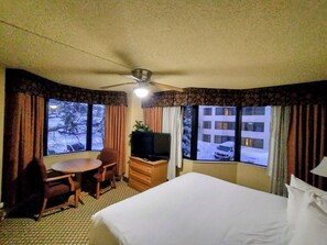 Une chambre d’hôtel comprenant un lit, un bureau, une chaise, une télévision et des fenêtres donnant sur un immeuble et des arbres.