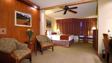 Habitación de hotel con cama, dos sillones, una mesita con un florero y un ventilador de techo, además de una ventana con cortinas.