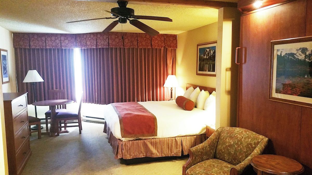 Ein Hotelzimmer mit einem Bett, einem Schreibtisch, einem Stuhl, einem Deckenventilator und einem Bild an der Wand.