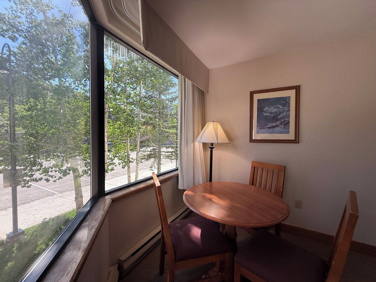 A hotel room with a dining table and chairs, a window with a view of trees, a lamp, and a framed picture on the wall.