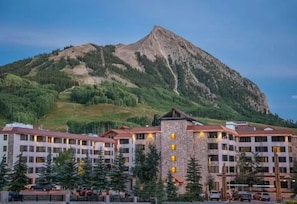 A hotel with multiple floors, a central tower, and a mountain backdrop. - NEW LISTING - 223 Slopeside 2 King Beds @ Base (Crested Butte)