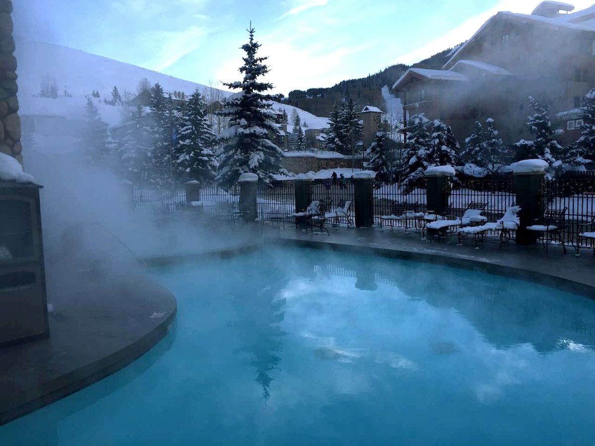 A pool with a clear blue water surface, surrounded by snow-covered trees and buildings.