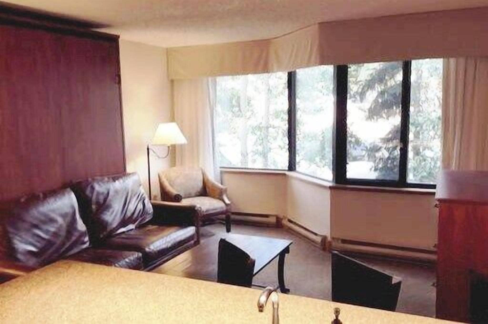 A room with a leather sofa, a small table, and a lamp. There are windows with curtains.