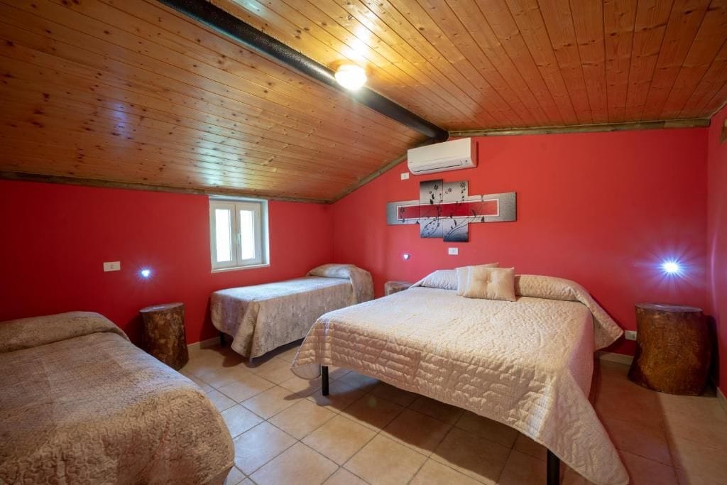 Une pièce avec un plafond en bois, un mur rouge et un lit recouvert d’une courtepointe. On y trouve une fenêtre et un climatiseur fixé au mur.
