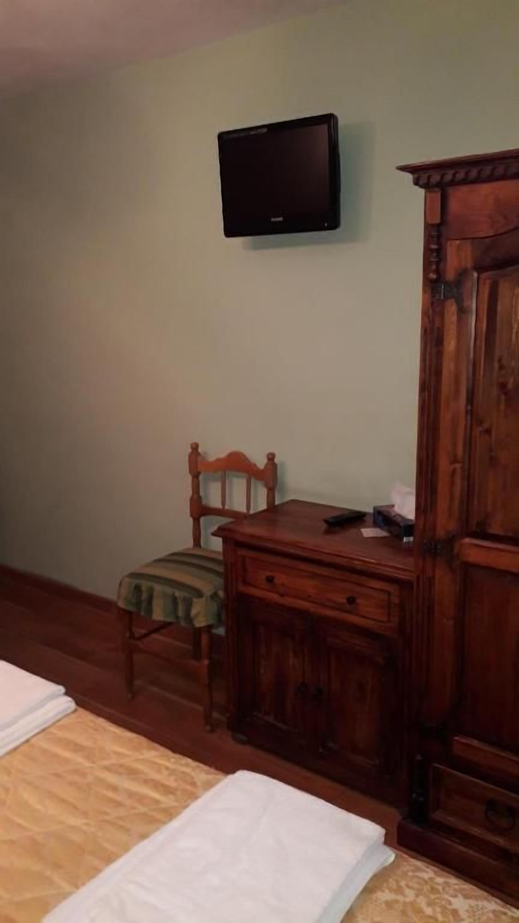A room with a bed, a wooden cabinet, a television mounted on the wall, and a chair with a cushion.