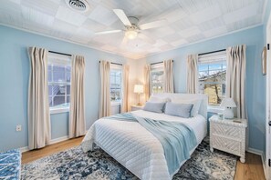 Un dormitorio con una cama, una mesita de noche, un ventilador de techo y una ventana con cortinas.