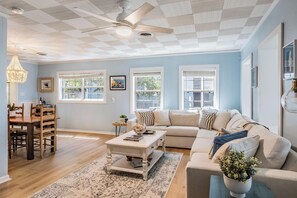 Ein Wohnzimmer mit einer Couch, Couchtisch und Essbereich mit Stühlen. Es gibt Fenster, einen Deckenventilator und eine Chandelier.