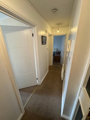 A hallway with a carpeted floor, white walls, and a door leading to a room. - Charming 1-bedroom Apartment in West Northamptonshire (Northampton)