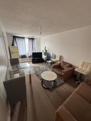 A compact living space with a glass dining table, a brown sofa, a television, and a small side table. - Charming 1-bedroom Apartment in West Northamptonshire (Northampton)
