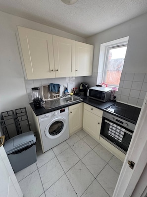A compact kitchen with a washing machine, microwave, and a small window. - Charming 1-bedroom Apartment in West Northamptonshire (Northampton)