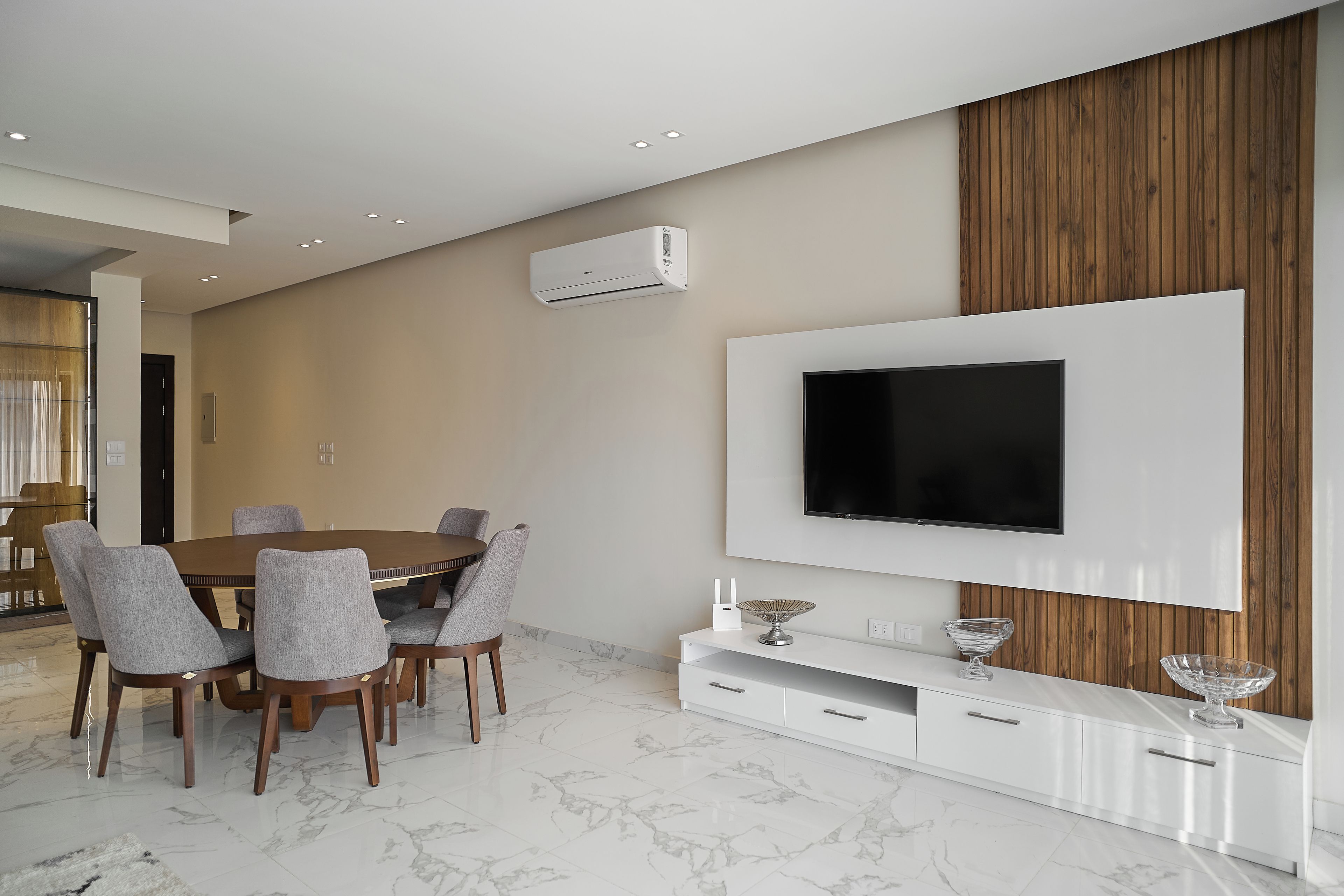 A modern dining area with a round table and chairs, a mounted flat-screen TV, and a white console with drawers.