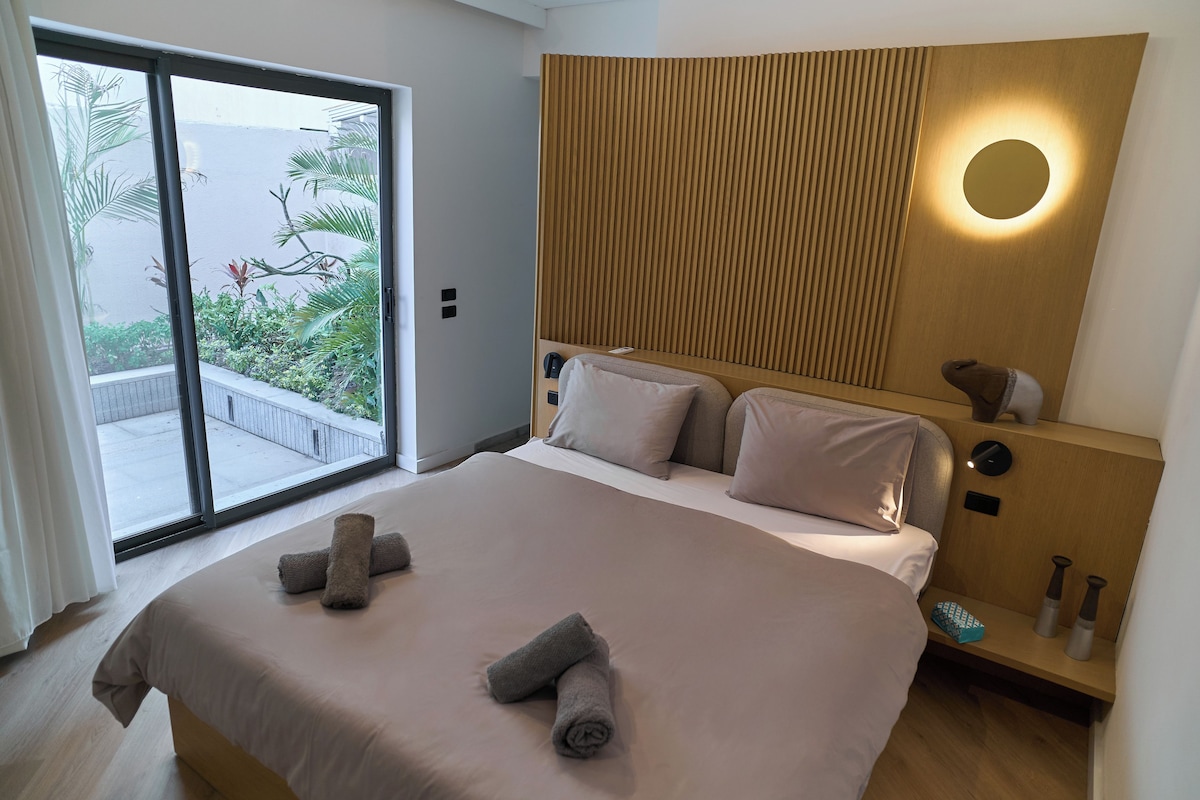 Habitación de hotel con una cama grande, una puerta corrediza de vidrio a un balcón y un cabecero de madera con una lámpara circular.