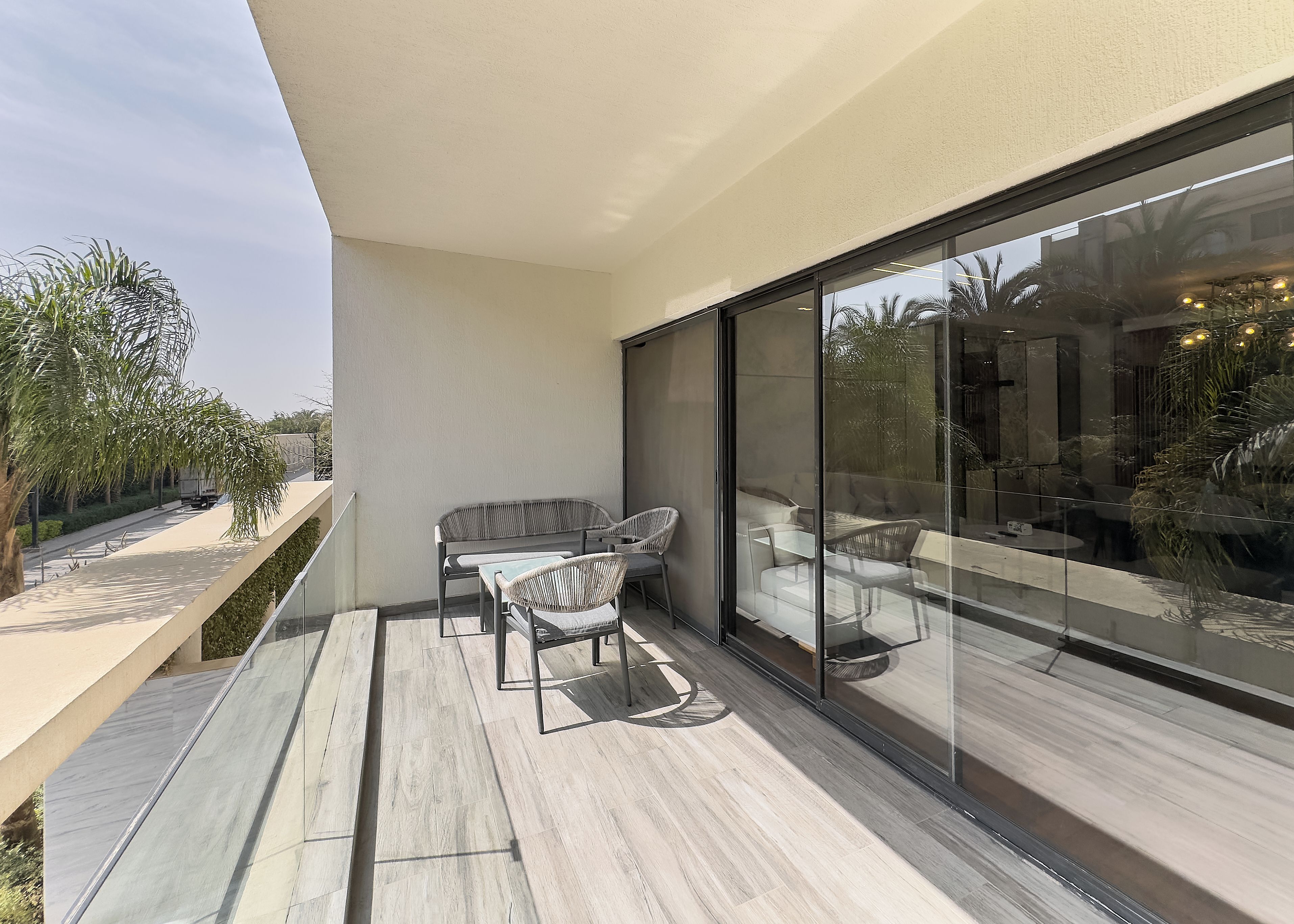 Un área de estar exterior moderna con una partición de vidrio, una terraza de madera y vistas a la calle y al verde.
