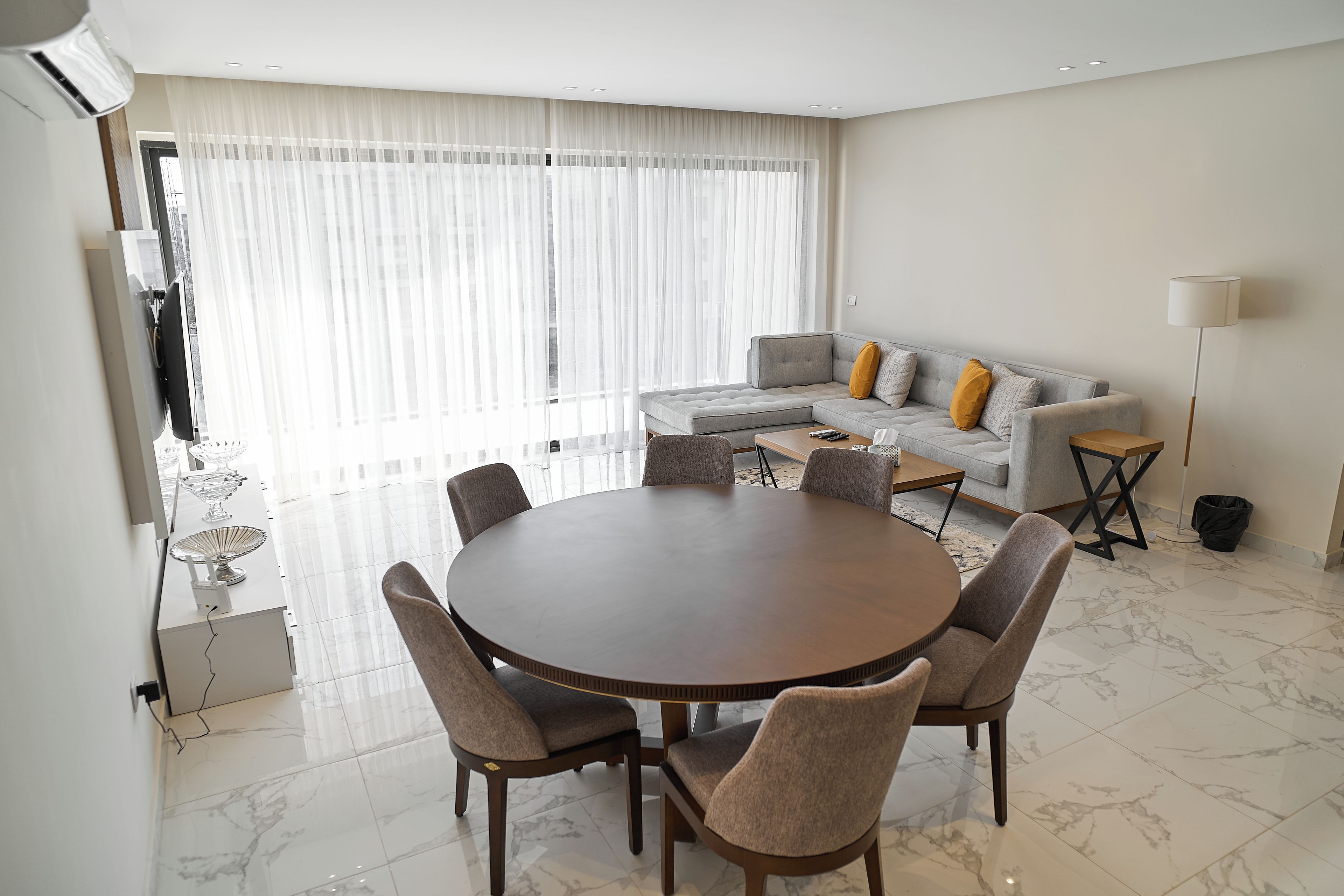 Un salón moderno con una mesa de comedor redonda, un sofá gris y un ventanal con cortinas traslúcidas.