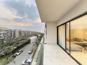 A balcony with a view of a residential street, a modern building, and a glimpse of a living room with a sofa. - Lakeview Residence Serviced By BlueKeys (New Cairo)