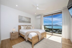 A bedroom with a bed, bedside table, lamp, and a large window with a view of the sea. - Ocean Views Private Villa (Townsville)