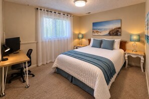 Ein Schlafzimmer mit einem Bett, einem Schreibtisch mit Computer, zwei Nachttischlampen und einem Wandbild mit Strandmotiv.