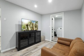 Ein Wohnzimmer mit einem Flachbildfernseher auf einem schwarzen Schrank, einem braunen Sofa und einem Durchgang zu einem Waschraum.