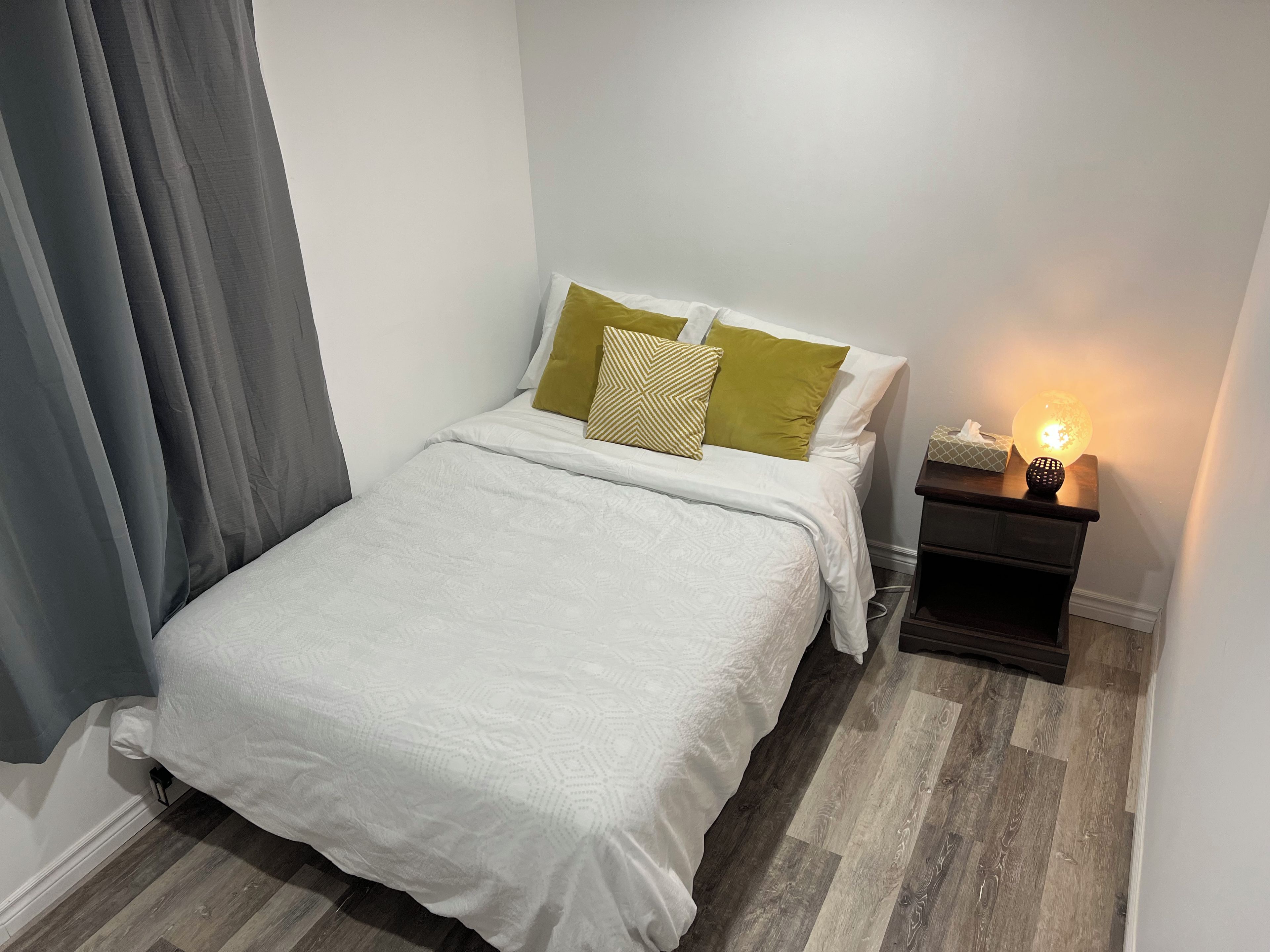 A neatly made bed with a white duvet, a bedside table with a lamp, and a wooden floor.