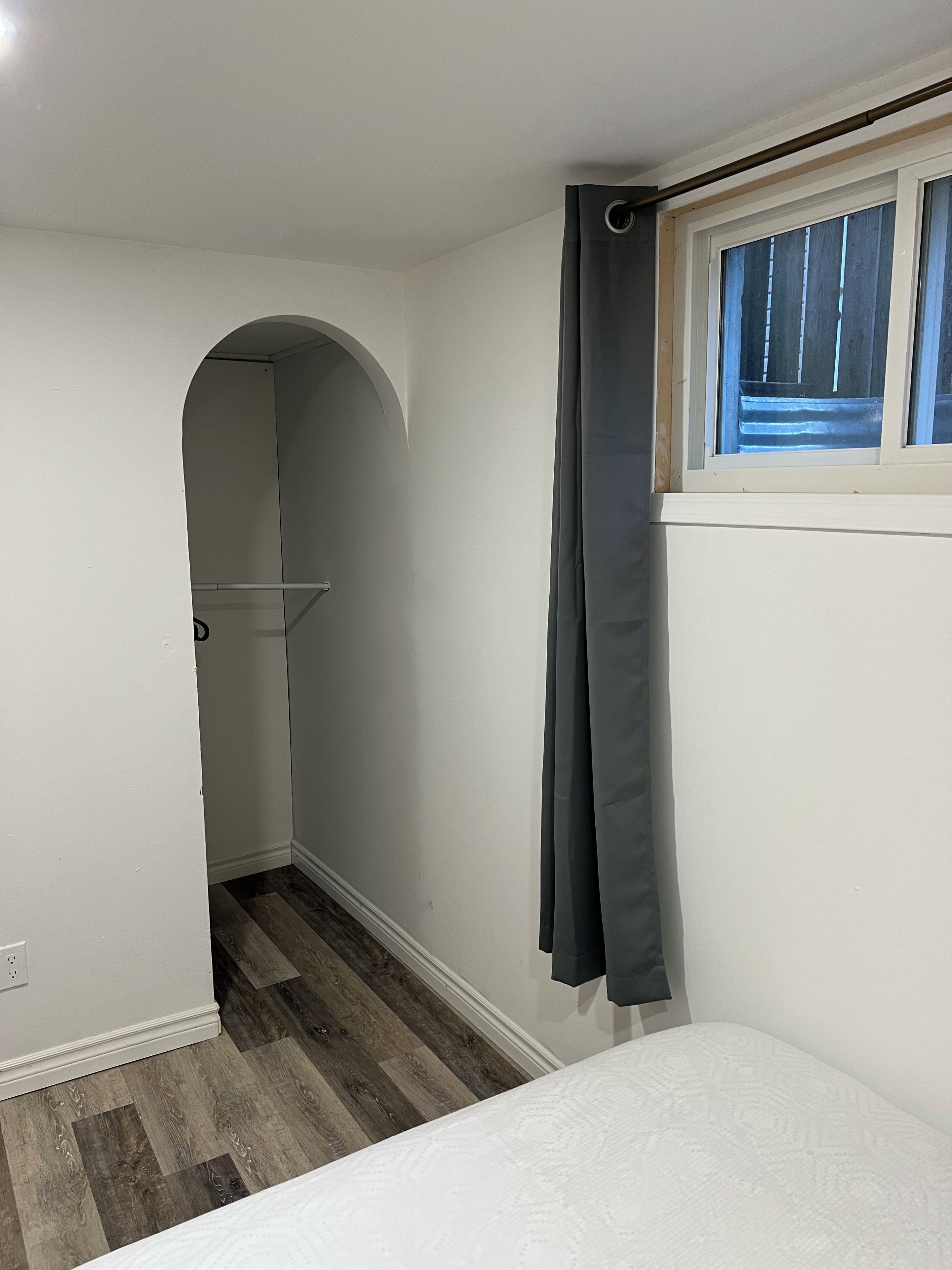 A narrow hallway with a door leading to a room, a window with a curtain, and a patterned bedspread.