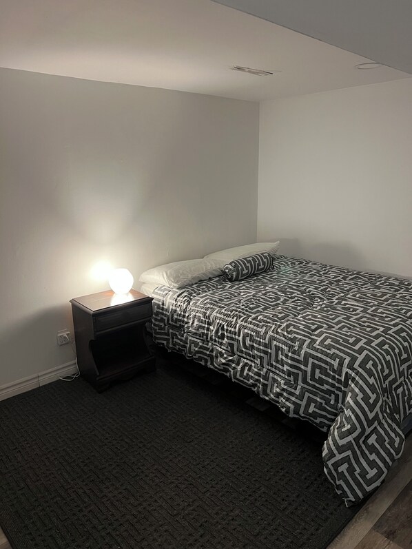 A bedroom with a bed, a nightstand with a lamp, and a carpeted floor.
