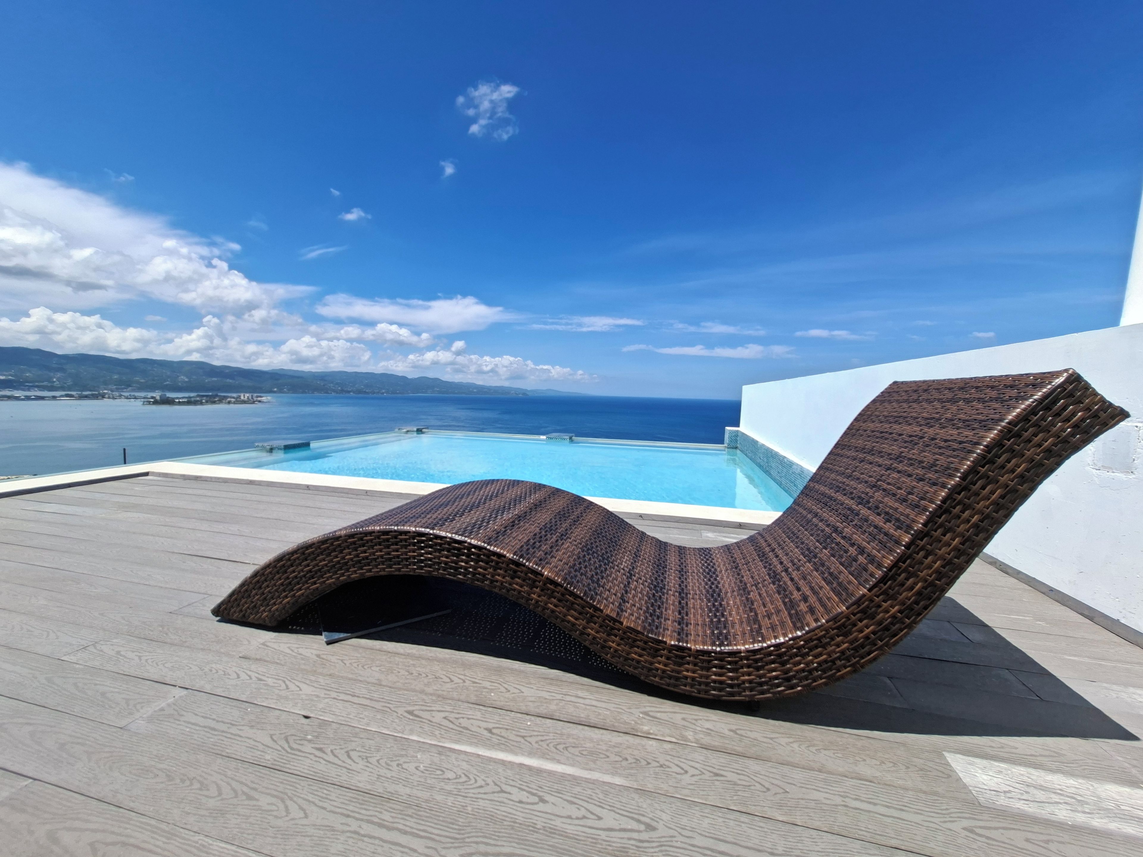 A wicker lounge chair on a wooden deck overlooking a pool with a view of the ocean and distant land.