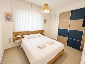 A hotel room with a bed, a bedside table, a lamp, a window with curtains, a wall-mounted TV, and a towel rack. - Port city haifa - Sea front 1 bdrm apt w/ mamad (Haifa)