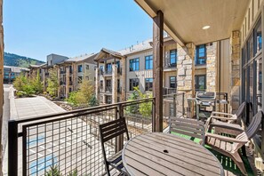 A balcony with a table and chairs, overlooking a courtyard with buildings and greenery. - Luxury 2BR Canyons Village Condo: Walk to Lift, Pool, Hot Tub & Viking Kitchen (Park City)