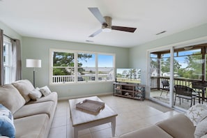 A spacious living room with a large window, a ceiling fan, and a view of the outdoors. - Bay Breeze Chincoteague | Bay Views + Modern Comfort! (Chincoteague Island)