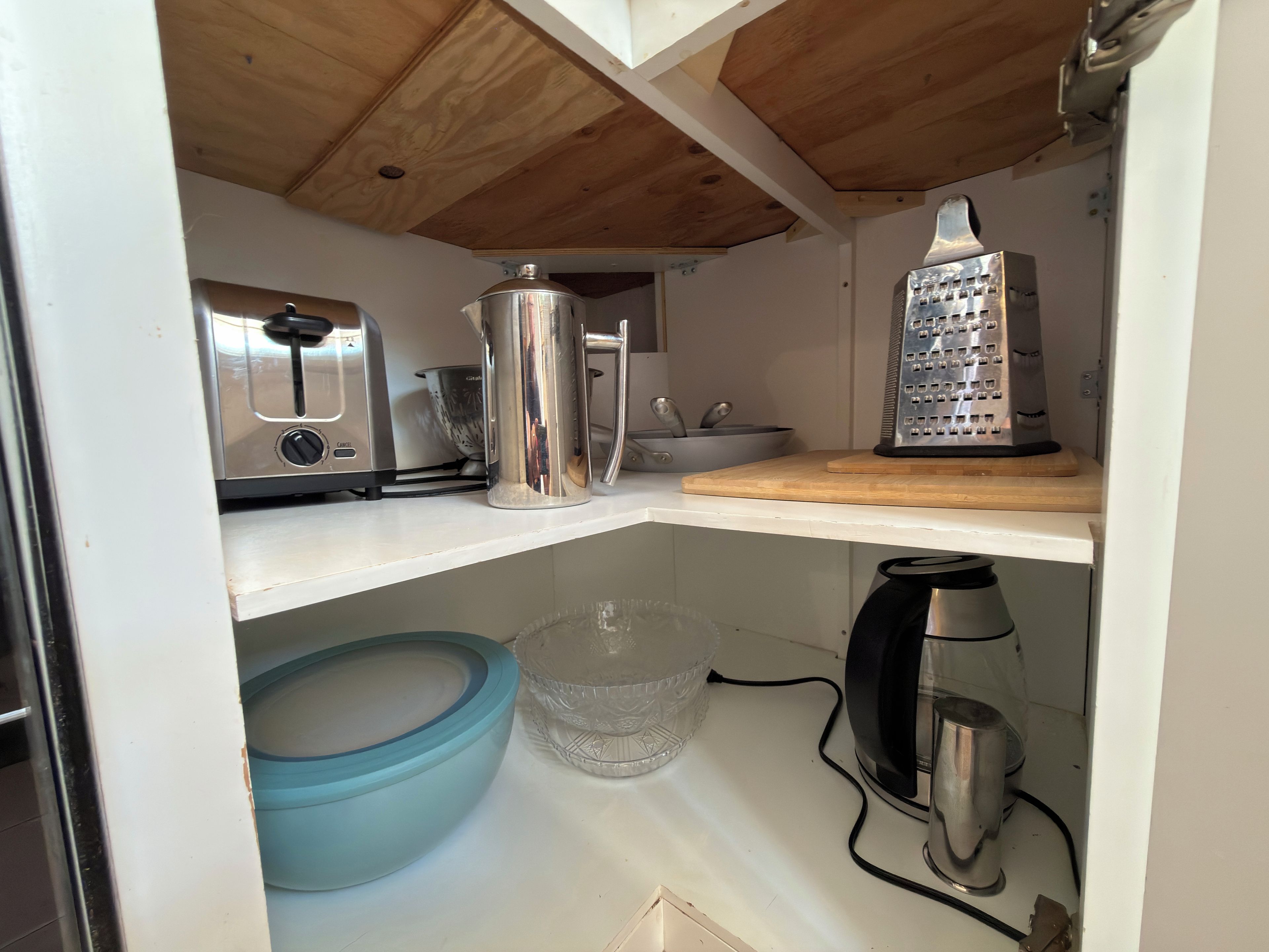 A compact kitchen with a toaster, kettle, and grater on shelves, a blue bowl, and a coffee maker on the counter.