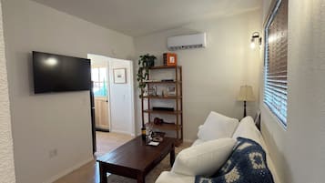 Una sala de estar con televisor de pantalla plana, una mesa de centro de madera, un sofá blanco con una manta azul, una estantería y un aire acondicionado instalado en la pared.