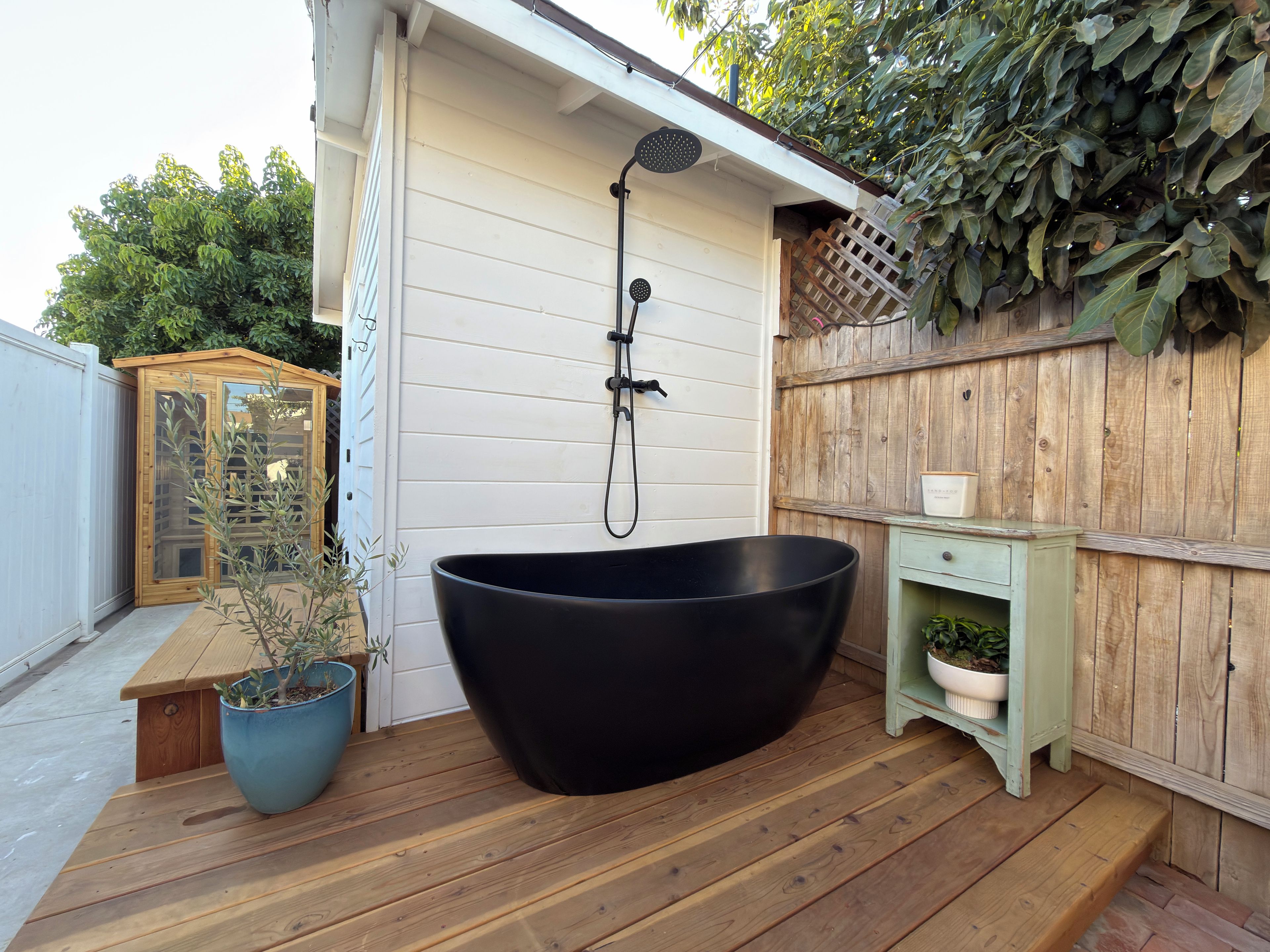Une baignoire noire sur une terrasse en bois, une clôture en bois et une plante en pot.