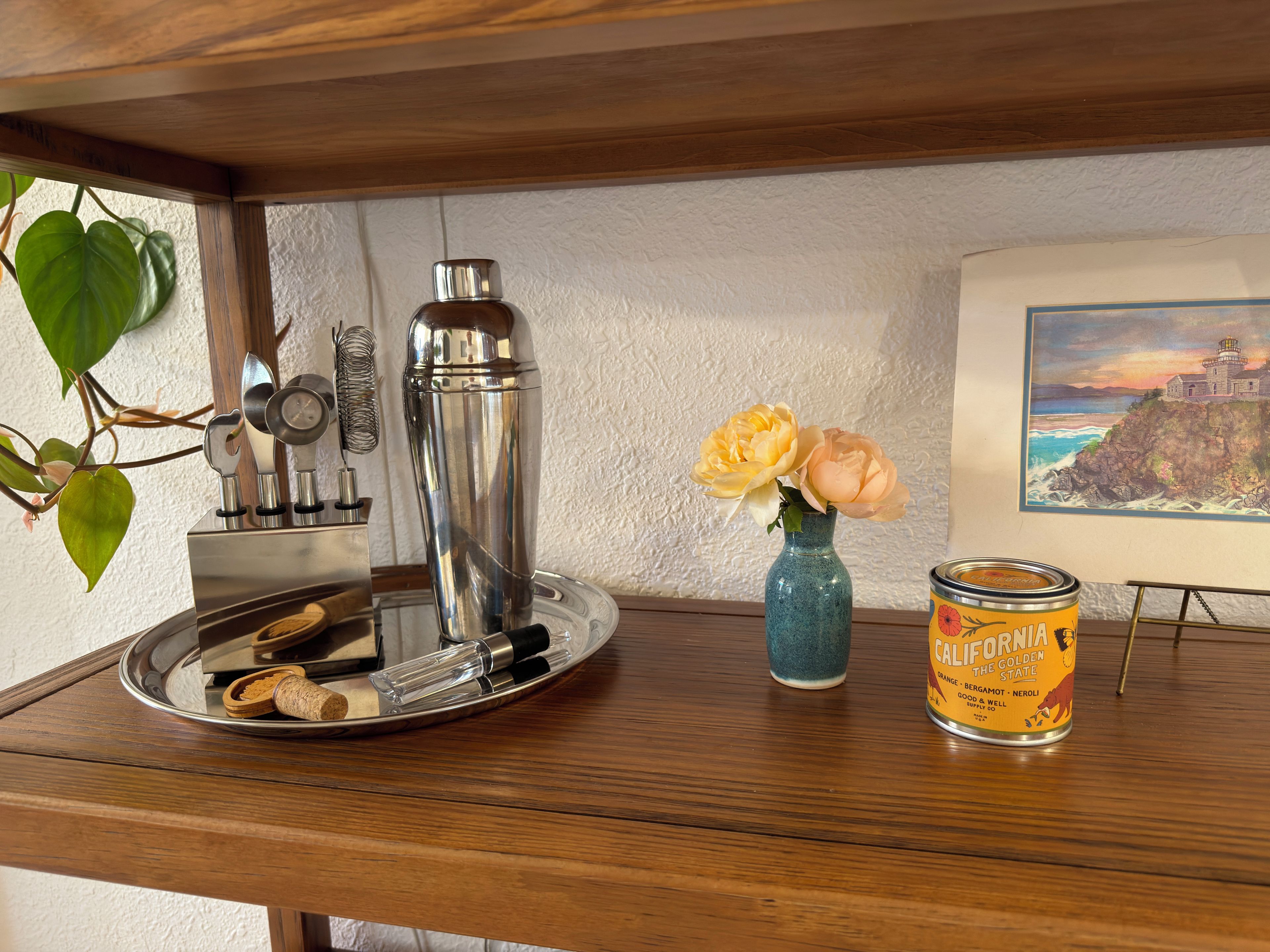 Una mesa de madera con un shaker de cócteles, un florero azul con flores y una lata de cerveza California Golden State.