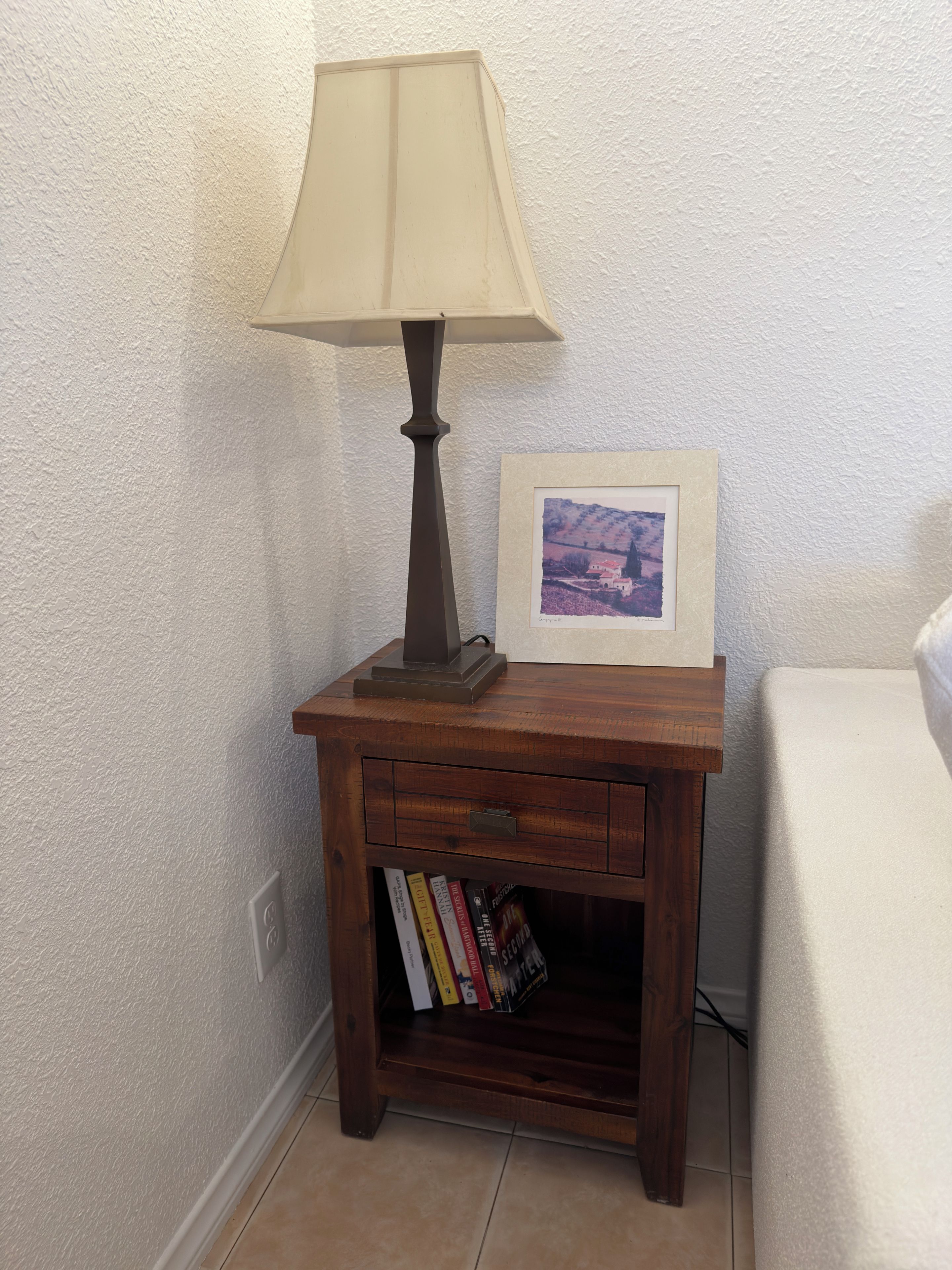A wooden nightstand with a lamp, a framed picture, and a book shelf.