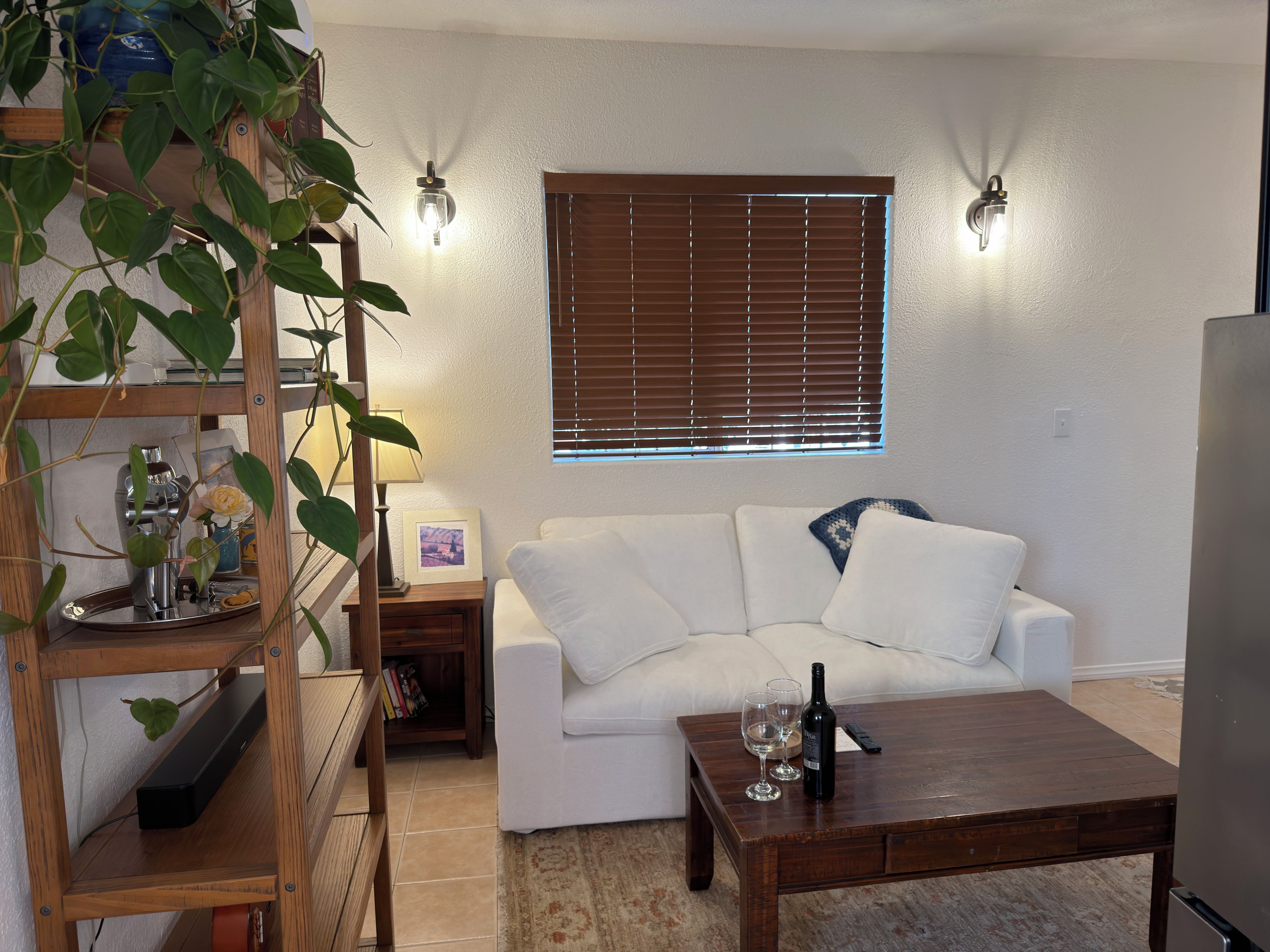 Una sala de estar con un sofá blanco, una mesa de centro de madera con copas de vino, un televisor y una ventana con persianas.