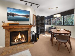 Een woonkamer met een open haard, een aan de muur bevestigde televisie, een eethoek met een tafel en stoelen, en een groot raam met uitzicht op bomen en een gebouw.