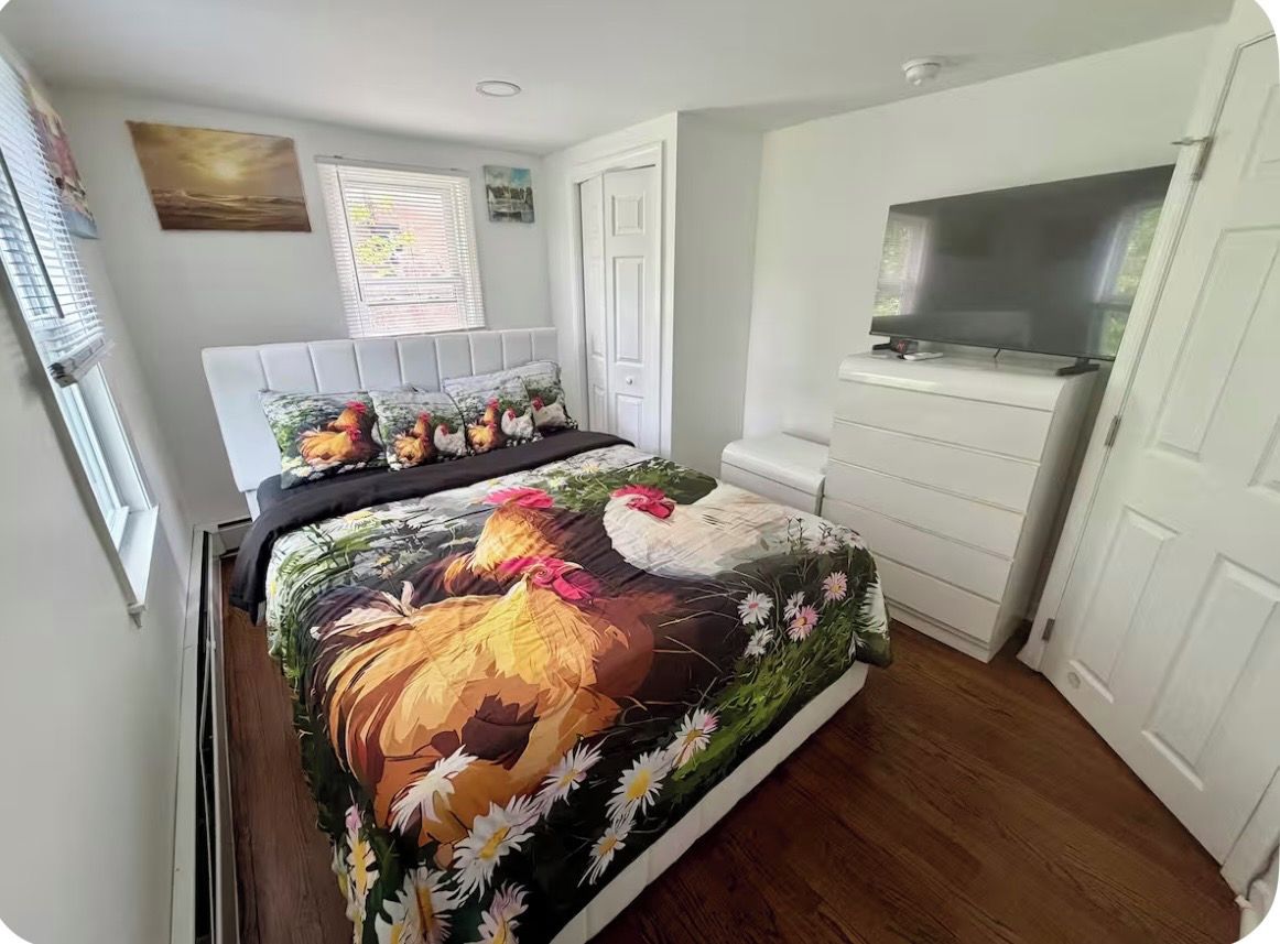 A bedroom with a bed, a TV, a white wardrobe, and a window with blinds.